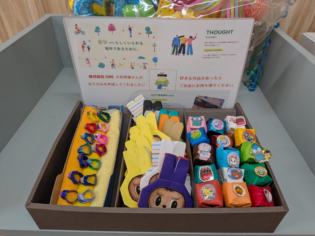 Tray filled with colorful origami and small wrapped gifts in front of a poster with Japanese text and illustrations.